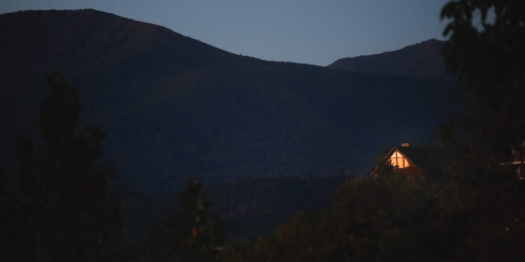 Minimalist photography of a cabin at dusk with illuminated window, framed by dark mountain landscape – photo by thefadedlens.com