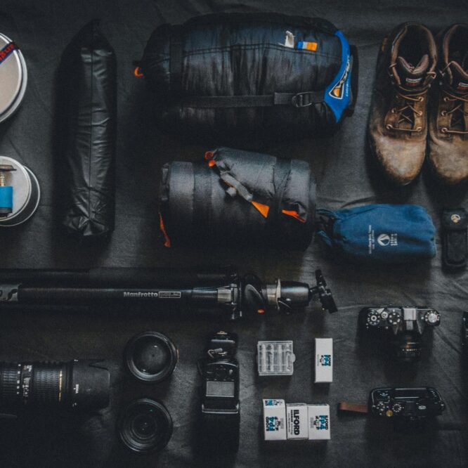 Flat lay of underrated photography accessories and outdoor gear, including camera bodies, lenses, Manfrotto tripod, film, sleeping bag, boots, and utility tools—perfect for adventure photographers.