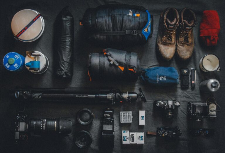 Flat lay of underrated photography accessories and outdoor gear, including camera bodies, lenses, Manfrotto tripod, film, sleeping bag, boots, and utility tools—perfect for adventure photographers.